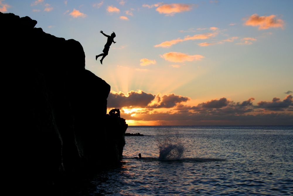 Boy jumps off a cliff into the ocean at Waimea Bay in Hawaii at sunset.