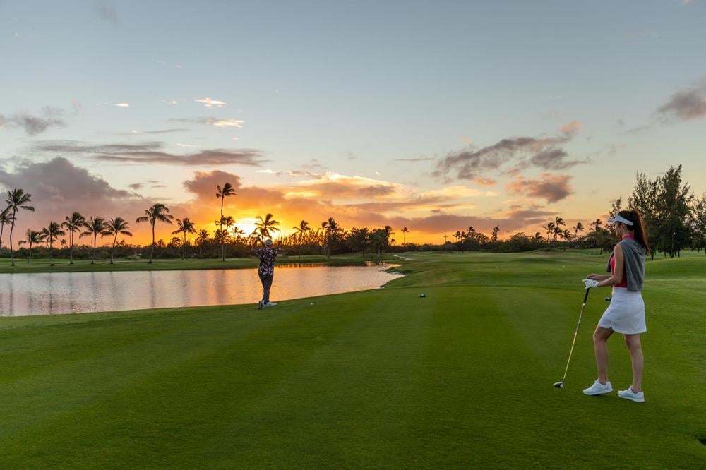 A woman overlooking a man golfing at the Hawaii Prince Golf Club during sunset.