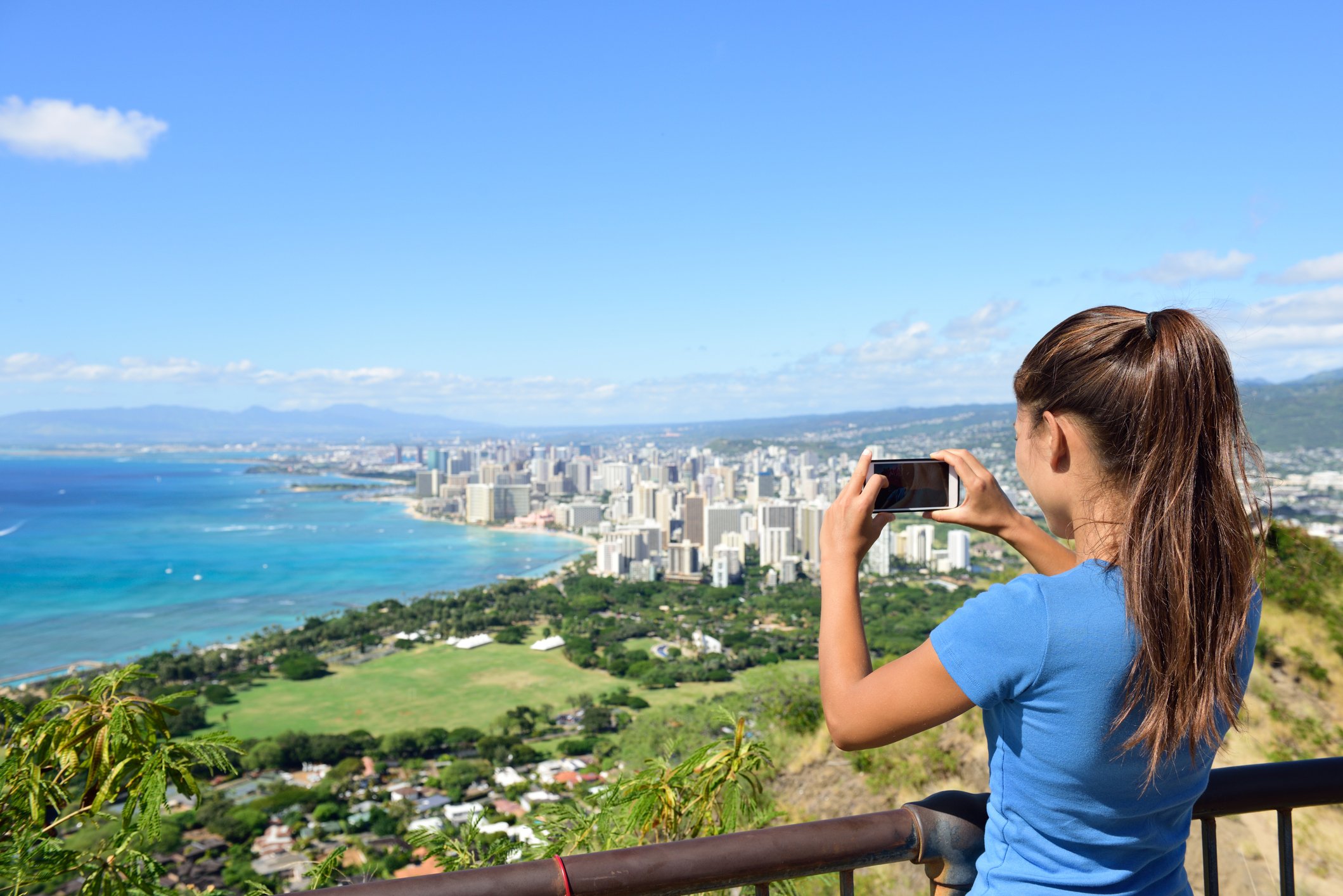 Hawaii tourist taking photo of Honolulu and Waikiki beach using smartphone camera. Woman tourist on hike visiting famous viewpoint lookout in Diamond Head State Monument and park, Oahu, Hawaii, USA.