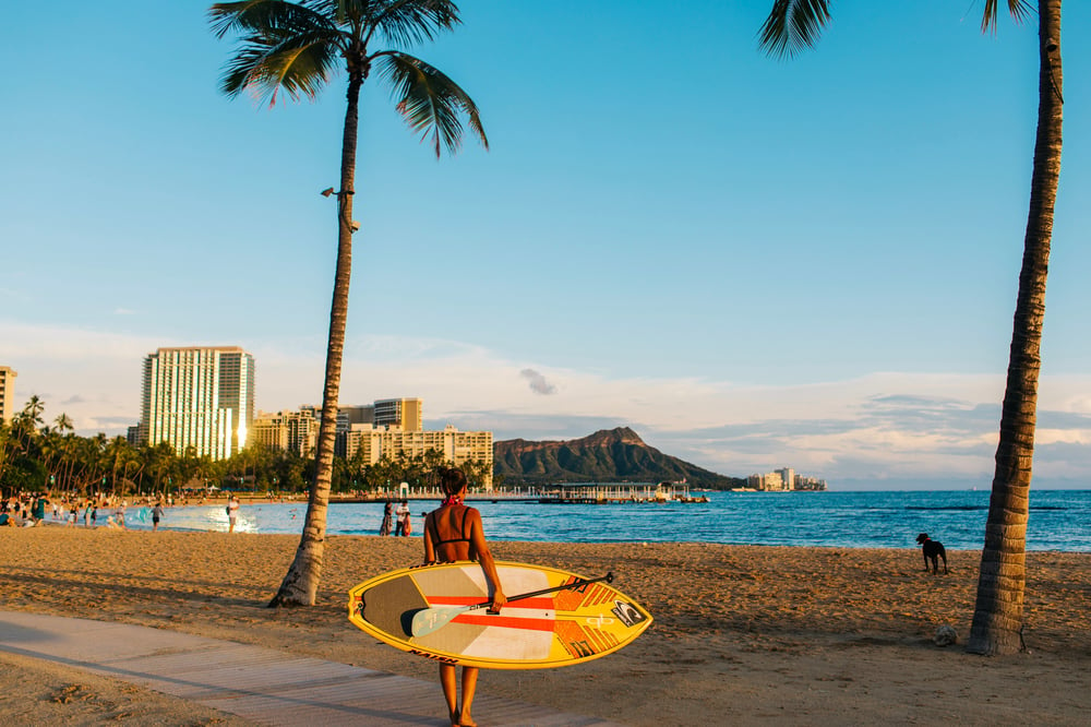 A woman with a surfboard walks toward Waikiki Beach on a sunny day