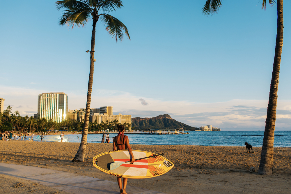 A woman with a surfboard walks toward Waikiki Beach on a sunny day