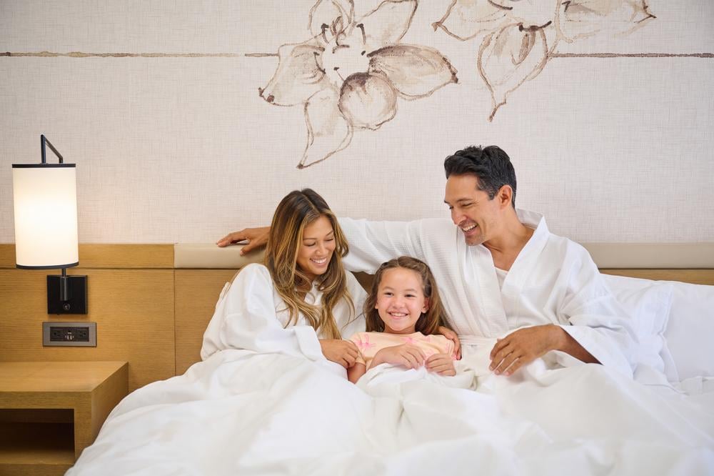 A family of three, mother, daughter and father cuddled up in bed at Prince Waikiki.