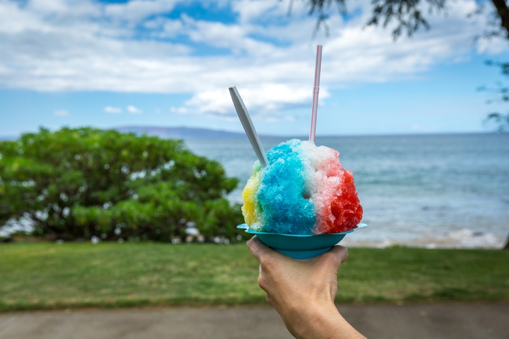 A hand holding a shave ice overlooking a beach in Honolulu, Hawaii.