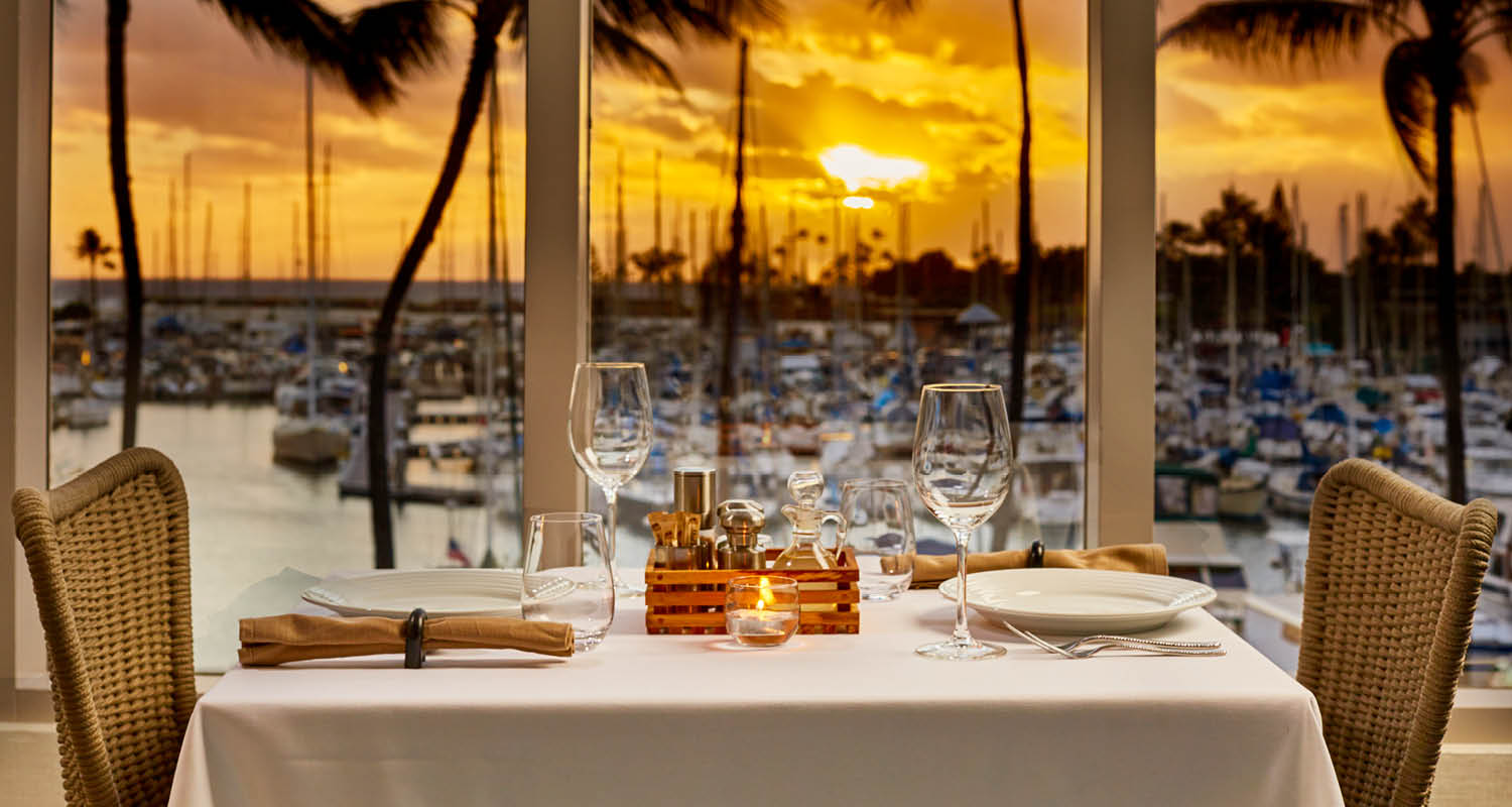 A table set for two with the sunset in view from the floor-to-ceiling windows at 100 Sails Restaurant & Bar in the Prince Waikiki Hotel.
