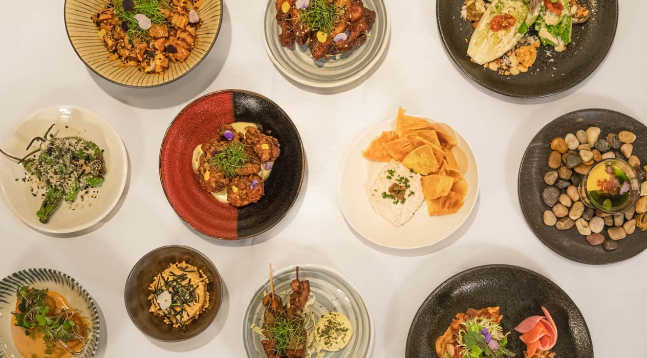 Plates of the 100 Sails Savory Menu displayed with Hurricane Tots, Chicken Wings, Romaine Salad, Oyster Shooter, Smoked Ahi Dip, Chicken Wings, Crab & Lobster Coquette and Kimchi Potato Salad.