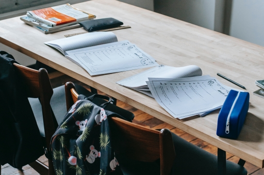 A classroom in Honolulu, HI with a table for two. Backpacks are placed on the back of the two chairs and school work with pencil cases are placed on the desk.